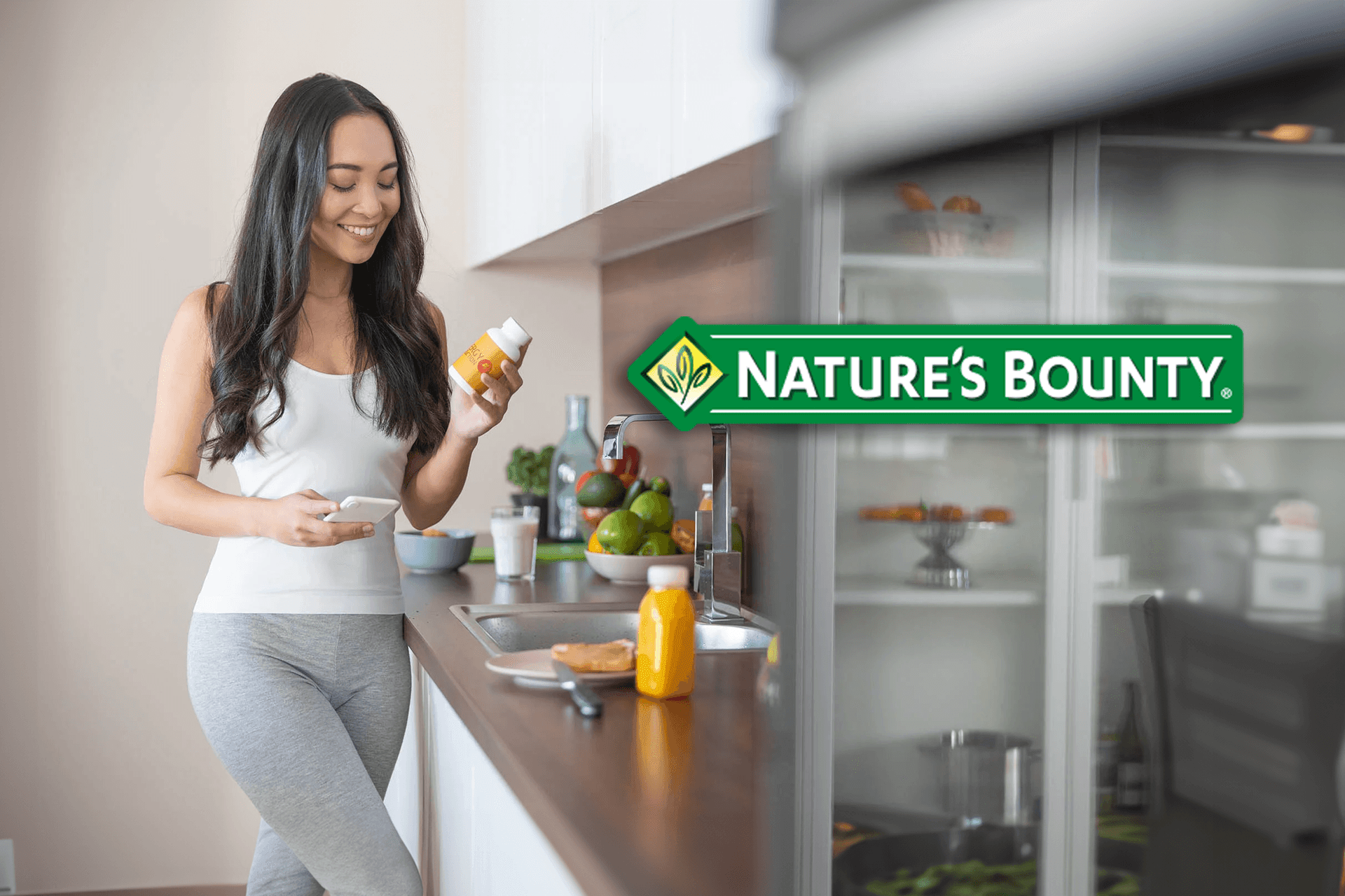 Person in athletic wear standing in kitchen holding Nature's Bounty supplement bottle and smartphone.