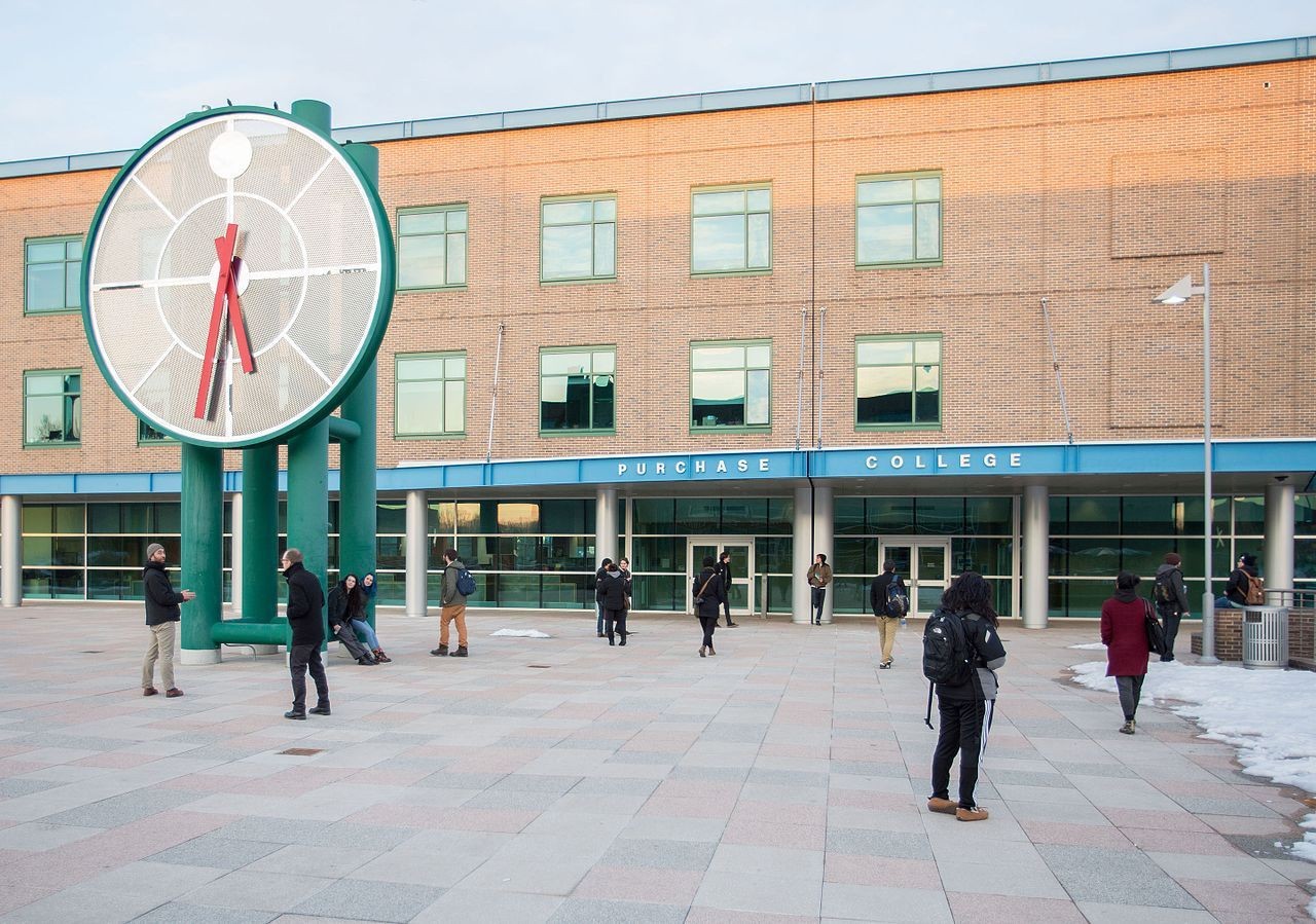 Students walking near a large outdoor clock in front of a building with 'Purchase College' sign.